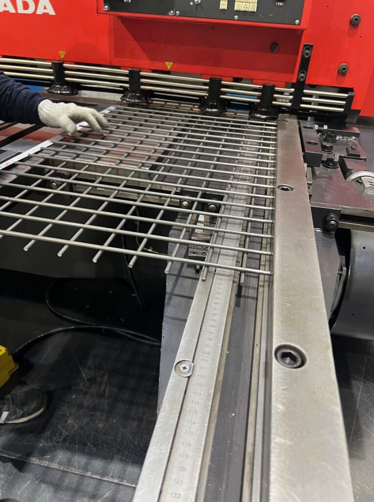 溶接金網の製造工程。作業員が機械にセットされた金網の寸法を、定規（スケール）を使って確認している様子。
