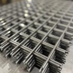 太い金属線が垂直・水平に交差し、強固に溶接された正方形メッシュの金網が、工場内で何枚も積み重ねられているクローズアップ写真。