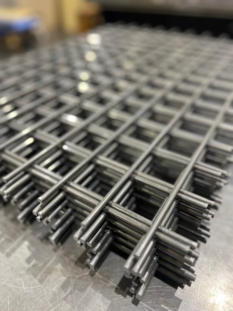 太い金属線が垂直・水平に交差し、強固に溶接された正方形メッシュの金網が、工場内で何枚も積み重ねられているクローズアップ写真。