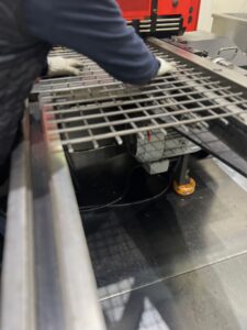 溶接金網を機械にセットし、作業の準備をする作業員のクローズアップ。手袋をはめた手が金網の端を調整しており、金網の下には機械のモーター部分が見える。