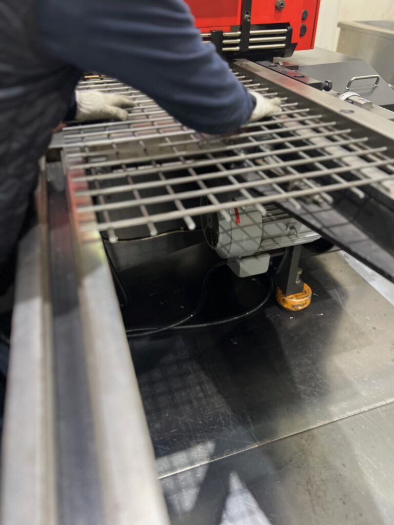 溶接金網を機械にセットし、作業の準備をする作業員のクローズアップ。手袋をはめた手が金網の端を調整しており、金網の下には機械のモーター部分が見える。