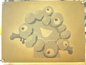 蓚酸アルマイト加工が施された金色の長方形のパンチングメタル板。板に開けられたドットのパターンが、2025年大阪・関西万博の公式キャラクター「ミャクミャク」の姿を表現している。