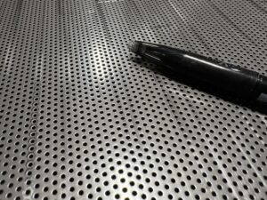 SUS304パンチングメタルの表面をクローズアップ。ペン先を影として映し、穴の小ささを強調している。